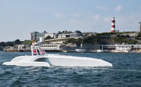 Hvidt autonomt skib, the Mayflower, sejler nær en kystlinje med en fyrtårn og bygninger i baggrunden