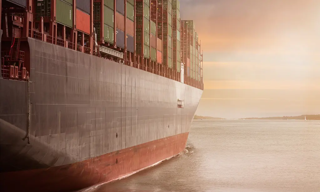 A container vessel at sea during a calm, golden sunset, with stacked containers visible along its hull.