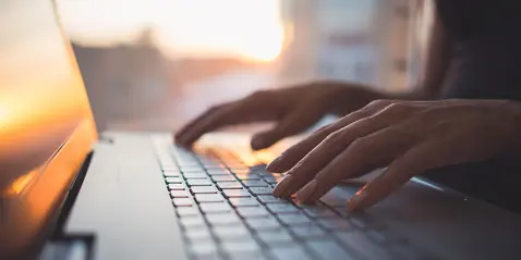Nærbillede af hænder, der skriver på en bærbar computers tastatur, med en solnedgang reflekteret på skærmen og en sløret baggrund.