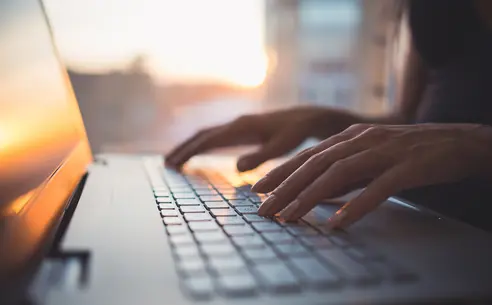 Close-up of hands typing on a laptop keyboard, with a sunset reflected on the screen and a blurred background.