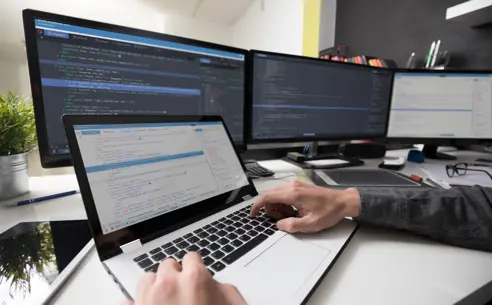 A person typing on a laptop with multiple screens showing code in the background
