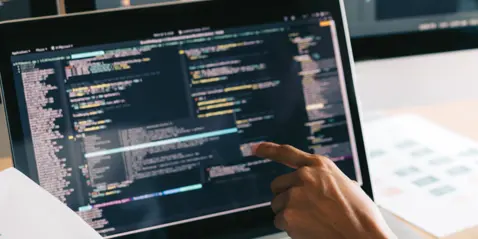 A person pointing at code on a laptop screen with multiple monitors in the background.