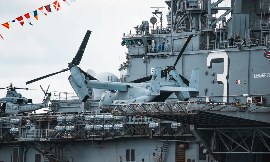 Militærhelikoptere på dækket af et stort marineskib, med farverige flag ovenover.