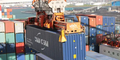 A crane lifting a blue container at a busy port with stacked containers.