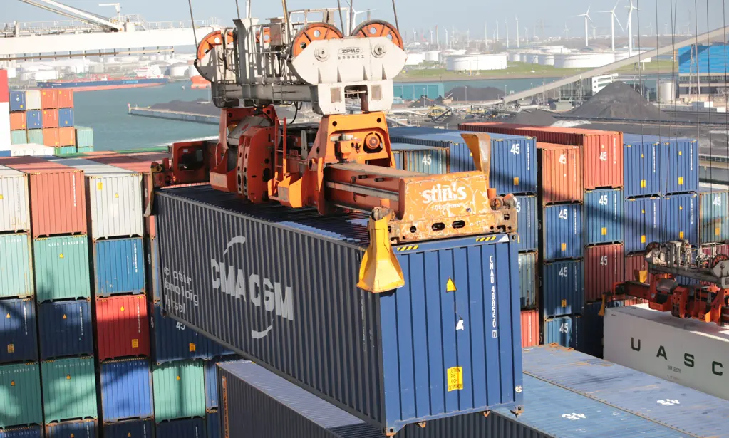 A crane lifting a blue container at a busy port with stacked containers.