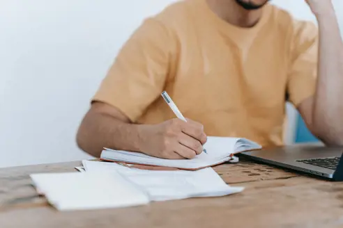 person, der laver noter på papir, mens han studerer på bærbar computer
