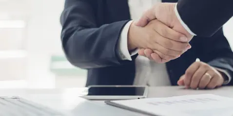 Handshake between two people in suits at a desk, with a laptop and documents in the foreground.