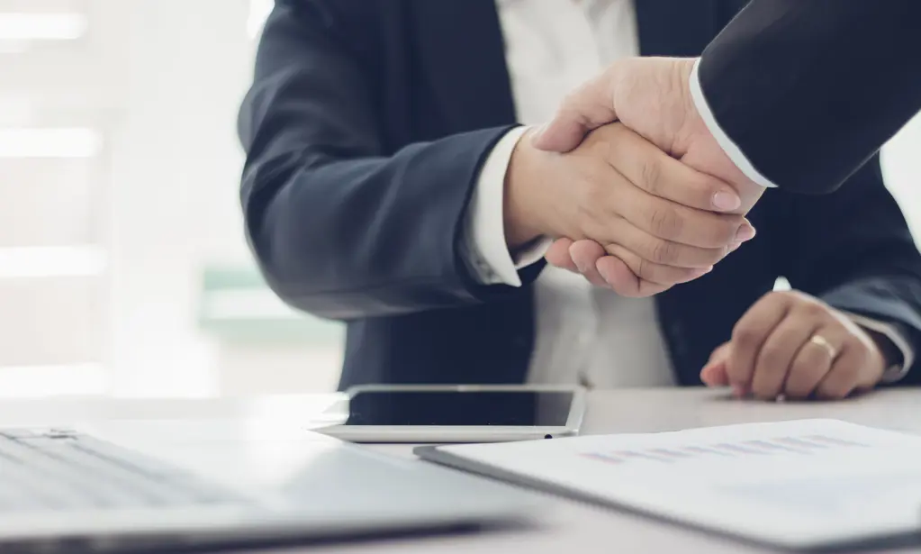Handshake between two people in suits at a desk, with a laptop and documents in the foreground.
