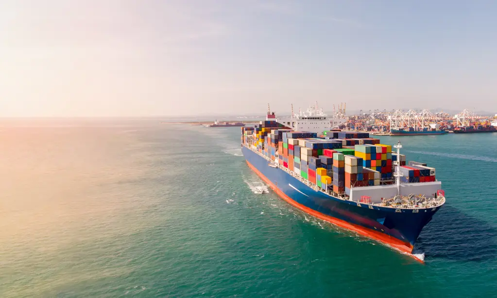 A container ship approaching a busy port filled with cranes and other ships.