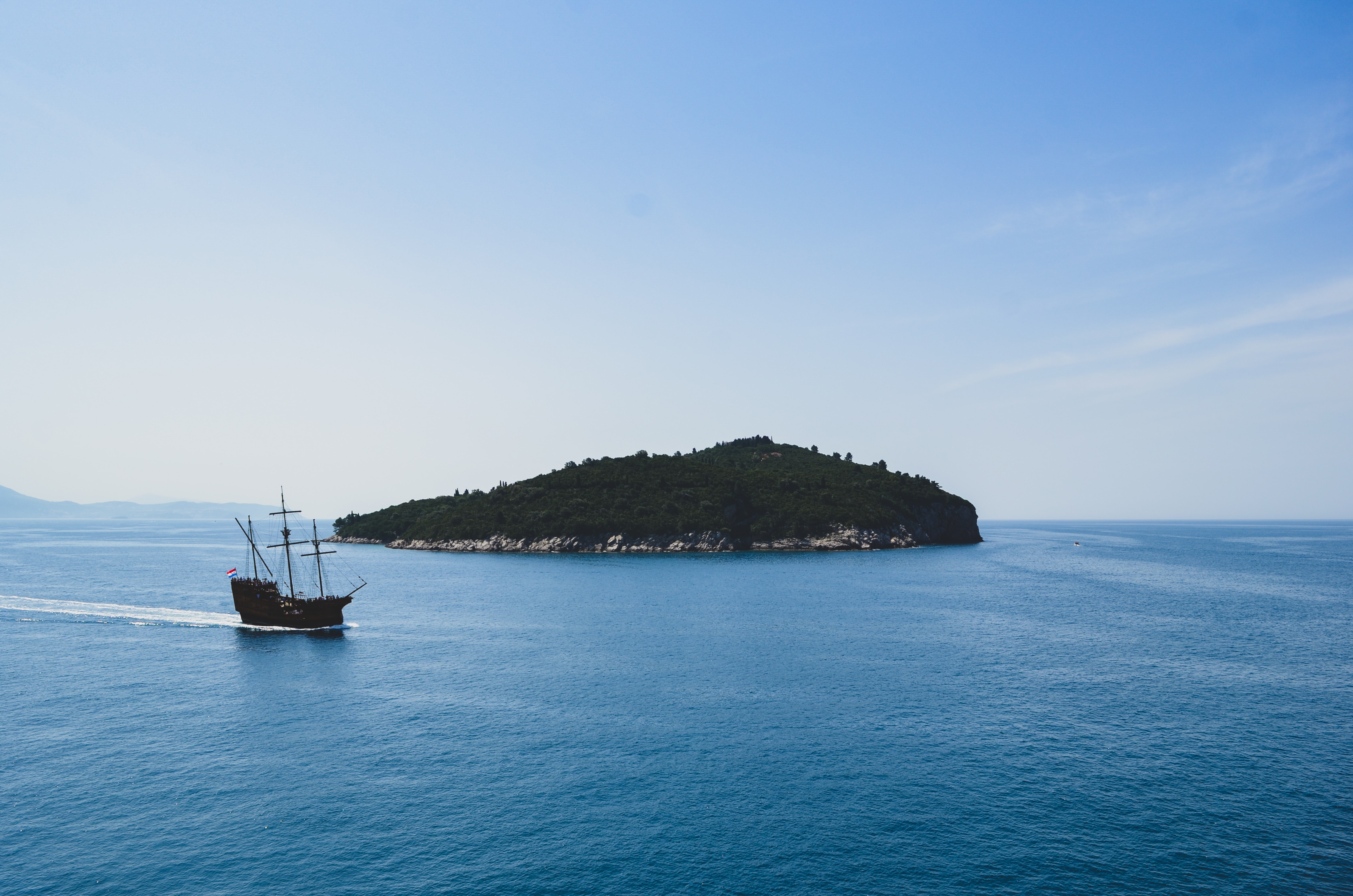 A ship passes by a small, green island surrounded by calm, blue waters under a clear sky.