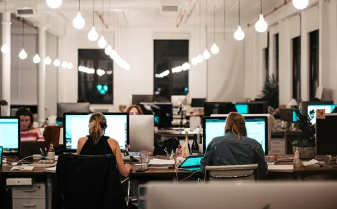 An open-plan office with people working at desks with computers, and hanging lights above.