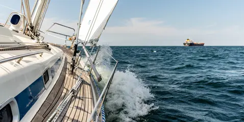 A sailboat cutting through waves with a large tanker in the distance on a clear day.